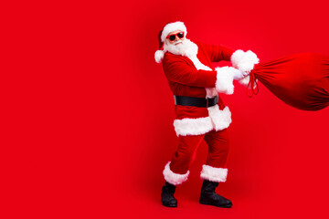 Full size photo of funky grandfather wear santa costume throwing bag with presents to empty space isolated on red color background.