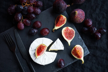 Ripe figs, red grapes with Camembert cheese next to cutlery on a dark background