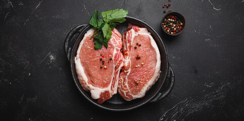 Cut raw meat pork steaks with seasonings in black cast iron pan, dark rustic stone background top view, ready for roasting. Pork loin chops cooking - obrazy, fototapety, plakaty
