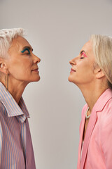 stylish senior female friends in blue and pink clothes standing with closed eyes on grey, side view