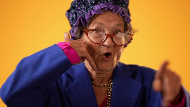 Closeup funny portrait of smiling happy elderly senior old woman with wrinkled skin smiling pointing to camera and giving call me gesture with hands isolated on solid yellow background.