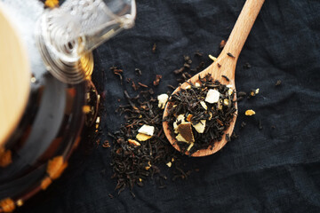 Black tea with pieces of fruit on a wooden spoon next to the teapot on a dark background