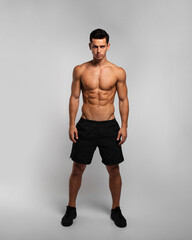 Full length image of a man with an athletic and fit body, posing in the studio with a bare torso, showing six abs pack, looking at camera,  grey background.