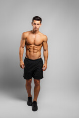 Full length image of a man with an athletic and fit body, posing in the studio with a bare torso, showing six abs pack, looking at camera,  grey background.