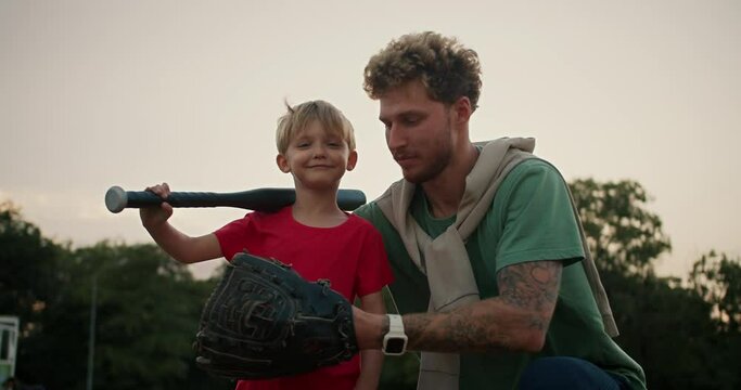Happy little blond boy in a red T-shirt holds a black baseball bat in his hands and throws a baseball into his dad's baseball glove in a Green T-shirt. Dad and son playing baseball in the park