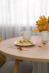Autumn home interior. Breakfast of cottage cheese with peach, ceramic vase with autumn leaves, candle, glass of coffee on round wooden table..