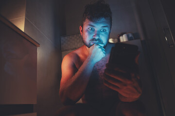 Phone light on face at night. Pensive guy using his phone in the toilet. Phone addiction.