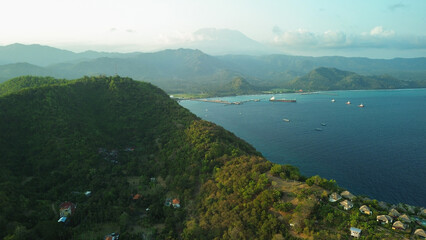 Aerial view of a hill illuminated by golden sunset light at Padang Bai port on the tropical island of Bali. View of sunken ships and the hotel where tourists relax.