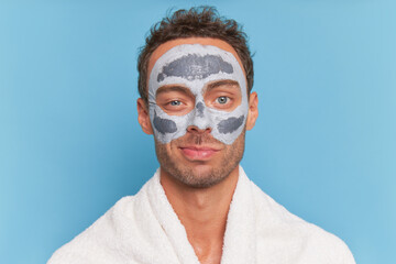 Close-up shot of young man with cosmetic clay mask on his face on blue background, male skincare concept, copy space