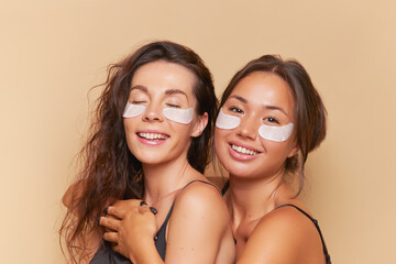 Two young women with cosmetic under eyes patches on their faces having fun while shooting in studio, skincare concept, copy space