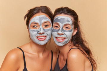 Two women with cosmetic clay masks on their faces having relaxing spa procedure with smiles, healthcare product concept, copy space