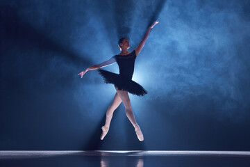 Elegant, artistic, tender ballerina, young woman dancing in tutu on stage over spotlight. Blue light. Concept of classical dance, art and grace, beauty, choreography, inspiration