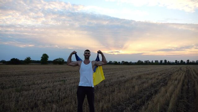 Ukrainian sportsman stand on barley meadow showing biceps with blue-yellow banner in hands. Young guy raised flag of Ukraine above head on wheat field at sunset. Victory against russian aggression