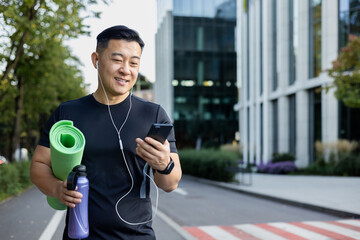 Young Asian male athlete in headphones goes to training on the city street. He holds a mat, a...