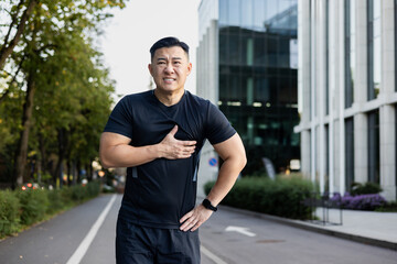 Portrait of an Asian young male sportsman standing on a city street and holding his chest with his hand, feeling severe pain, overload, looking tiredly at the camera