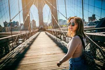 Girl in sunglasses strolls along the Brooklyn Bridge in New York City at sunset.