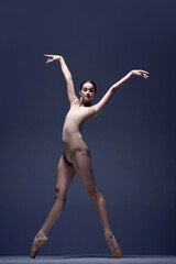 Beautiful, slim, elegant woman, ballerina in motion, posing in beige bodysuit and pointe against blue studio background. Concept of classical dance, art and grace, beauty, choreography, inspiration