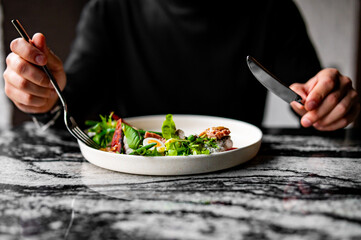 Hands of a man with a fork and knife eat Salad nicoise. Tuna, with green beans, tomatoes, potatoes and a vinaigrette mustard dressing in white plate