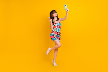 Full length photo of shiny excited woman dressed flower print outfit showing v-sign tacking selfie modern device isolated yellow color background