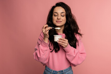 Waist-up portrait of lady eating the yogurt with the teaspoon from the plastic cup in front of the...