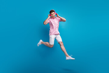 Full length portrait of carefree overjoyed person jumping hands touch headphones isolated on blue color background