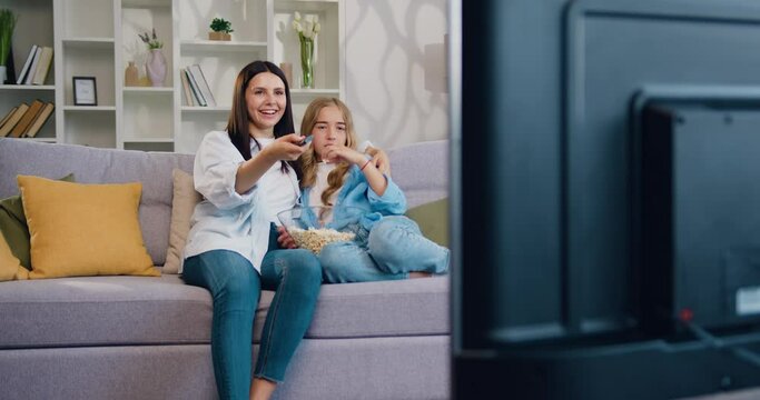 Cute teen daughter and smiling mother laughing sitting on couch eating popcorn enjoy funny television show. Woman with children girl sitting on sofa at home watching TV with popcorn together. Happy