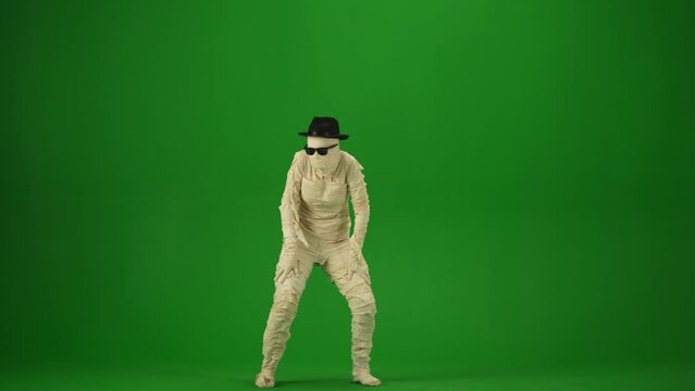 Scary mummy wrapped in bandages dances wearing a black hat and sunglasses. Green screen isolated chroma key. Mock up, workspace for your promotion clip or advertisement. Full length.