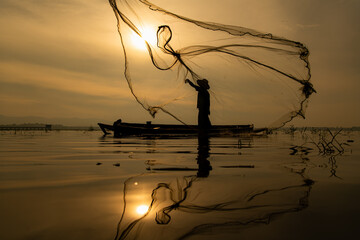 Silhouette of fisherman at sunrise, Standing aboard a rowing boat and casting a net to catch fish...