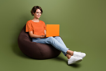 Full length photo of smart girl dressed t-shirt in eyewear sit on bean bag typing look at laptop...