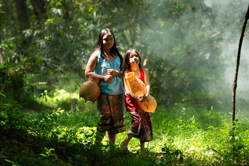 Two girls Asian women with traditional clothing stand in the rainforest. They had fun playing together before assisting Grandpa in catching fish in a nearby lake. - obrazy, fototapety, plakaty