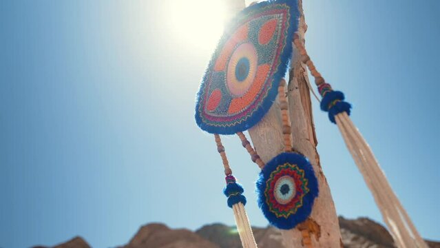 Close up knitted evil eye amulet hangs on a wooden branch stick in the desert at sunset. Blue sky and mountains in the background. Spirituality and shamanism in the wild nature