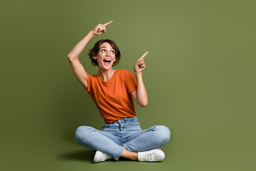 Full length photo of impressed girl dressed t-shirt sit on floor look directing at promo empty space isolated on green color background - obrazy, fototapety, plakaty