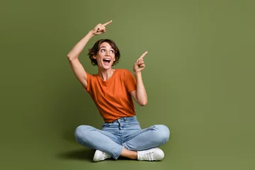 Tuinposter Full length photo of impressed girl dressed t-shirt sit on floor look directing at promo empty space isolated on green color background © deagreez