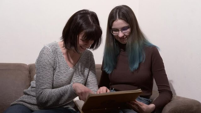 Mother and daughter watch family photos, remember pleasant moments. Beautiful woman and her teenage daughter are looking through photos and smiling while sitting on couch at home.