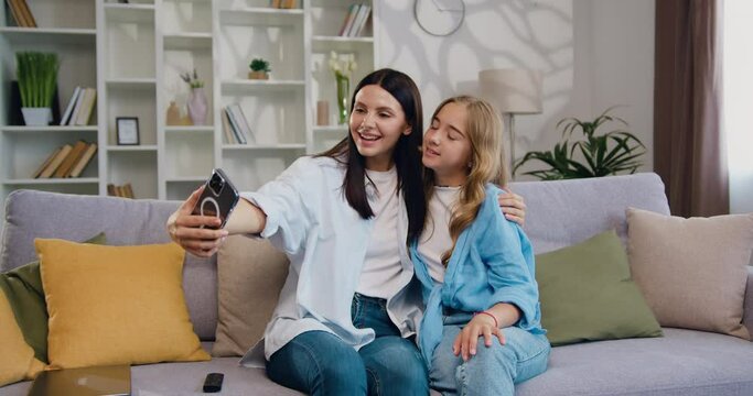 Cheerful girl and mother parents while taking funny picture on a phone at home. Beautiful smiling family make selfie pictures on smartphone camera on sofa. Having fun and relaxing over the weekend.