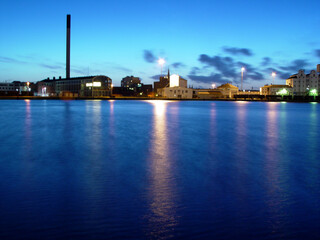 Sea, night and buildings in port with lights, reflection on calm blue water or holiday location. City at ocean in evening, travel, skyline and nature, urban beach village for vacation, peace and view