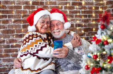 Smiling senior couple with Santa hats hugging enjoying Christmas time together video chatting with...