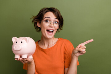 Photo of surprised young funny girl holding piggy bank directing finger novelty promoter invest...