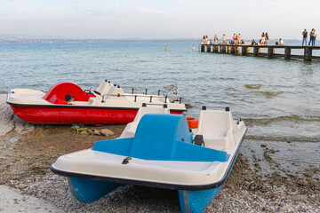 a red pedal boat and a blue pedal boat tied on the beach of a lake. In the background a pier with some people - 664002996