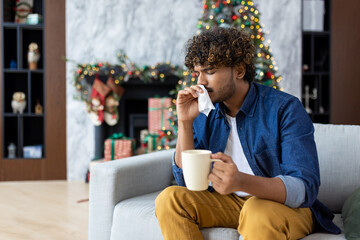 Sick man sitting at home alone on Christmas, holding a cup of hot drink and a napkin, hispanic cold...