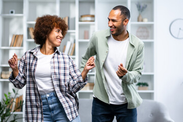 Cheerful woman and man dressed in casual wear laughing and playfully dancing together at home
