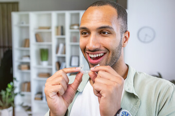 Young man holding dental aligner at home with a happy face standing and smiling with a confident smile showing teeth - obrazy, fototapety, plakaty