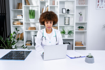 Young lady doctor in white medical uniform with stethoscope using computer laptop talking video...