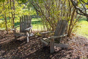 Landscape view of a pair of wooden adirondack chairs in garden setting with dappled sunlight

