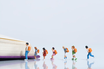 Schoolchildren with schoolbags stand in a row in front of an open book, miniature figures scene, white background, copy space - obrazy, fototapety, plakaty