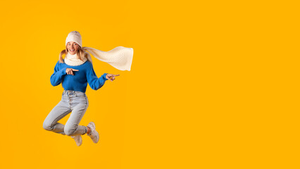 Obraz premium Excited caucasian woman in knitted hat jumping and pointing aside at free space on yellow studio background