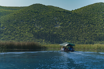 Podróż po jeziorze. Góry jezioro i łódka. Turyści na łodzi. Ludzie w łódce na jeziorze wśród gór. Ludzie i góry. Jezioro i ludzie. Wysokie góry w Chorwacji. Delta Neretwy - obrazy, fototapety, plakaty