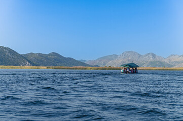 Podróż po jeziorze. Góry jezioro i łódka. Turyści na łodzi. Ludzie w łódce na jeziorze wśród gór. Ludzie i góry. Jezioro i ludzie. Wysokie góry w Chorwacji. Delta Neretwy - obrazy, fototapety, plakaty