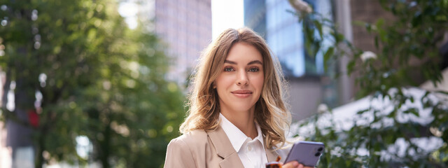 Close up portrait of corporate woman, young intern walks on street to office, holds mobile phone, texts messages on smartphone