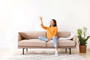 Happy young asian woman sitting on couch, turning on AC